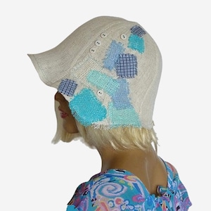 Può includere: Un cappello cloche in lino beige con toppe di tessuto blu e bianco cucite sopra. Il cappello ha un'ampia tesa e un dettaglio con bottoni.