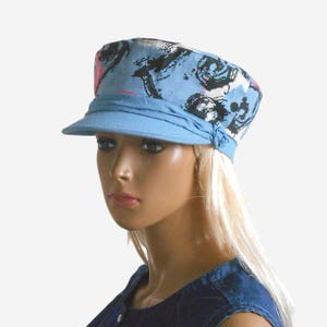 May include: A light blue military-style hat with a short brim. The hat features a fabric band and is decorated with a black, white, and pink abstract pattern. The hat is worn by a mannequin.