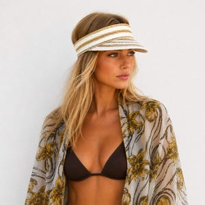 May include: A white and gold striped visor with a wide brim. The visor is worn by a person wearing a brown bikini top and a sheer, floral-patterned kimono. The kimono has a light gray background with gold and black floral designs.