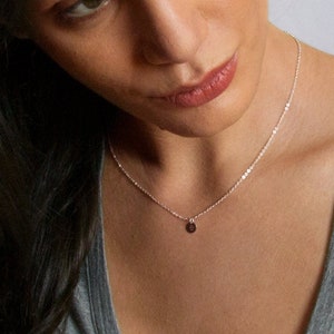 May include: A fine silver necklace with a small, circular pendant. The necklace is worn by a person in a grey top. The pendant is dark, and the chain is delicate and shimmering. The background is plain white.