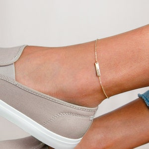 Peut inclure: Un bracelet de cheville doré avec un charm rectangulaire est porté sur une cheville. Le charm a un petit motif. La cheville est au-dessus d'une paire de baskets grises à enfiler avec des semelles blanches. L'arrière-plan est clair.