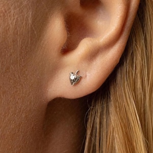 May include: A silver strawberry stud earring with a small, sparkling gem in the centre. The earring is worn on an ear, showcasing the delicate design and detail of the jewellery. The earring is a small, elegant piece of jewellery.