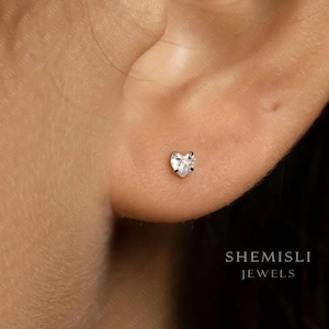 Peut inclure: Gros plan d'une boucle d'oreille en forme de cœur sertie d'un diamant. La boucle d'oreille est sertie dans un sertissage en argent ou en or blanc et est portée sur une oreille. Le nom de la marque "SHEMISLI JEWELS" est visible.