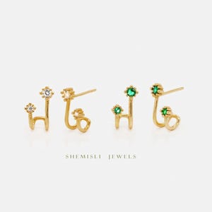 Peut inclure: Boucles d'oreilles dorées au design unique. Une paire présente des pierres claires et scintillantes, l'autre des gemmes vertes vibrantes. Les boucles d'oreilles ont une forme délicate et incurvée, avec une petite boucle en bas. Le texte "SHEMISLI JEWELS" est en bas.