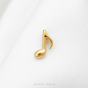 Peut inclure: Une petite breloque en forme de note de musique dorée avec une surface lisse et réfléchissante. Le design présente une tige unique et un crochet incurvé, reprenant la forme classique d'une croche. Le texte SHEMISLI JEWELS est inscrit en bas.