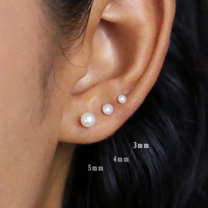 May include: Three white pearl stud earrings, sized 3mm, 4mm, and 5mm, are shown on an ear. The earrings are round and have a shiny finish. The ear is set against a neutral background.