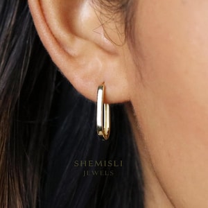 May include: A close-up of a gold-coloured, rectangular hoop earring. The earring is worn on an ear, with the brand name "SHEMISLI JEWELS" visible in the lower portion of the image. The earring has a polished, reflective surface.