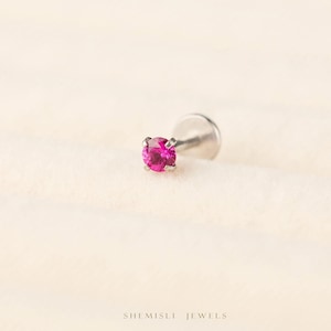 May include: A single silver stud earring featuring a vibrant pink gemstone. The round gemstone is held in place by four prongs. The earring is presented on a soft, cream-colored background. The brand name "SHEMISLI JEWELS" is visible at the bottom.