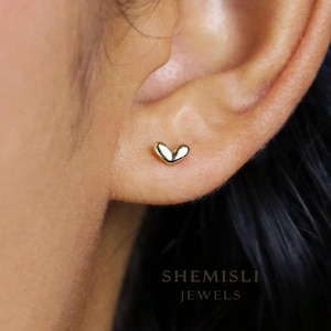 Peut inclure: Une petite boucle d'oreille en forme de cœur de couleur or. La boucle d'oreille est sur une oreille, avec le nom de la marque "SHEMISLI JEWELS" en dessous. La boucle d'oreille a une surface lisse et polie et un design simple et élégant.