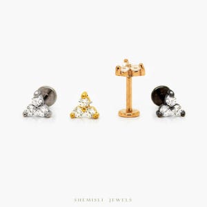 May include: Four pieces of jewellery are displayed against a white background. The jewellery includes three-stone stud earrings and a labret stud. The earrings and labret studs are in silver, gold, and black metal finishes, each with clear, sparkling stones.