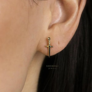May include: A gold-coloured sword-shaped earring. The earring is a stud style with a detailed hilt and blade. The earring is worn on the ear. The text "SHEMISLI JEWELS" is visible.