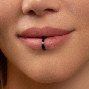 May include: A close-up shot of a person's lips, featuring a black lip ring. The ring is a plain, circular design. The skin around the lips is smooth and even-toned, and the lips are a natural, neutral colour.