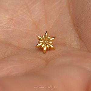 May include: A small, gold-coloured metal stud earring shaped like a six-pointed snowflake with faceted, pointed petals radiating from a central point. The piece features a polished, reflective surface and is positioned against a skin-toned background.