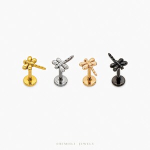 May include: Four dragonfly-shaped lip piercing studs in gold, silver, rose gold, and black. Each stud features a detailed dragonfly design atop a circular base. The studs are displayed against a white background. The brand name "SHEMISLI JEWELS" is at the bottom.