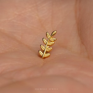 May include: A small gold-toned stud earring shaped like a delicate botanical leaf sprig with a central stem and six symmetrical leaves. The piece features a polished metallic finish and is positioned against a skin-toned background. Text reads SHEMISET JEWELS.