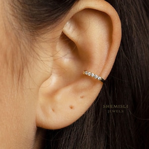 May include: A gold ear cuff featuring a line of small, clear gemstones. The ear cuff is worn on the upper ear. The text "SHEMISLI JEWELS" is visible in the lower right corner.