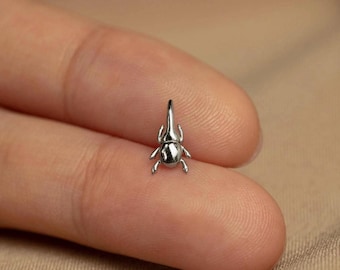 May include: A small, polished silver-toned metal charm shaped like a rhinoceros beetle. The charm features a smooth, reflective surface with a distinct horn and six legs, designed as a miniature insect figurine with a metallic finish.