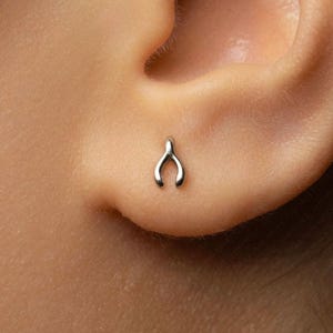 May include: A small, silver-toned wishbone-shaped stud earring positioned on an earlobe. The earring features a smooth, metallic finish with a Y-shaped design, consisting of two curved prongs meeting at a single point at the top of the piece.
