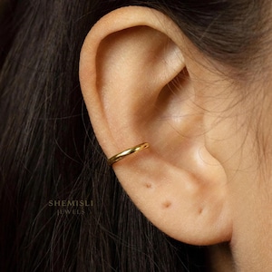 May include: A close-up of a gold ear cuff. The ear cuff is a simple, thin band that wraps around the ear cartilage. The ear is in focus, with the cuff's smooth surface reflecting light. The brand name "SHEMISLI JEWELS" is visible. A piece of jewellery.