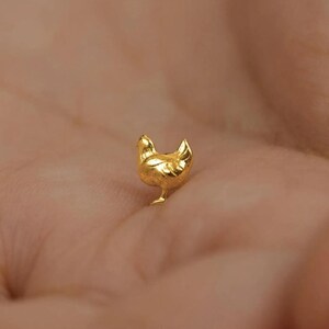May include: A small, gold-coloured chicken figurine. The miniature sculpture is detailed with a textured surface, showcasing the bird's form. The chicken is standing upright, with its head and tail feathers visible.