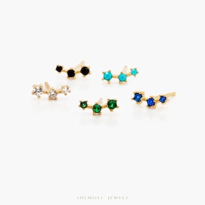 May include: Several gold-coloured stud earrings, each with three gemstones in black, turquoise, emerald green, sapphire blue, and clear. The earrings are displayed on a white surface. The brand name is visible.