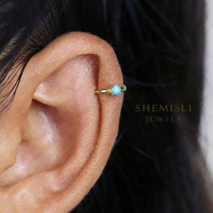 May include: A gold ear cuff featuring a light blue gemstone. The jewellery piece is designed to clip onto the ear cartilage. The text "SHEMISLI JEWELS" is visible in the background.