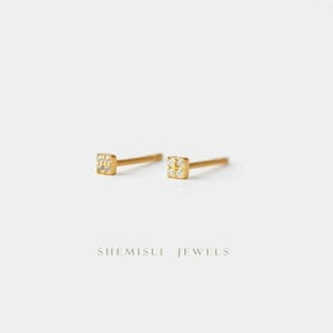 May include: Two gold-coloured square stud earrings, each featuring small, clear gemstones. The earrings are set against a white backdrop. The text "SHEMISLI JEWELS" is printed below the earrings.
