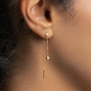 May include: Gold chain earrings featuring a small, round, clear gemstone at the top and a larger gemstone dangling below. A thin, rectangular gold bar hangs at the bottom of the chain. The earrings are worn in an ear piercing.