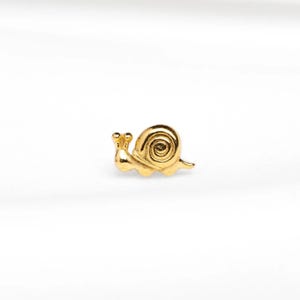 May include: A small, gold-coloured snail figurine. The snail has a detailed spiral shell and two antennae. The figurine is set against a white background.