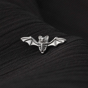 May include: A silver-coloured metal pin shaped like a bat with outstretched wings. The bat features a detailed skeletal chest design and pointed ears. The surface of the metal has a polished finish with recessed areas that highlight the bat's anatomy.