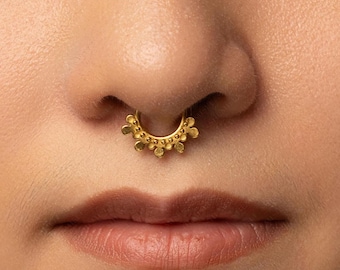 Könnte beinhalten: Ein goldener Septumring mit einem halbkreisförmigen Band, das entlang der Außenkante mit einer Reihe kleiner, erhabener runder Perlen verziert ist, wodurch ein wellenförmiges Aussehen entsteht. Der Schmuck sitzt mittig am Nasenseptum.