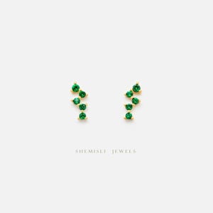 Peut inclure: Deux boucles d'oreilles dorées, chacune ornée de cinq pierres vertes rondes disposées en grappe. Les boucles d'oreilles sont présentées sur un fond blanc, avec le texte "SHEMISLI JEWELS" en dessous.