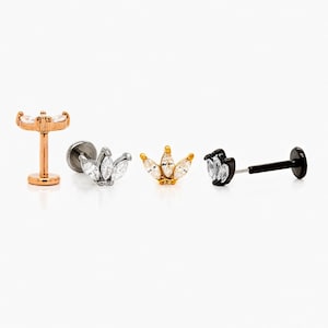 May include: Four pieces of body jewellery are displayed against a white background. The jewellery includes a rose gold labret stud with three clear marquise-cut stones, a silver labret stud with three clear marquise-cut stones, a gold labret stud with three clear marquise-cut stones, and a black labret stud with a clear marquise-cut stone.