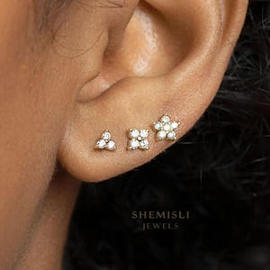 May include: Three gold earrings with clear gemstones are displayed. The earrings are shaped as a triangle, a square, and a flower. The brand name "SHEMISLI JEWELS" is printed in white at the bottom of the image.