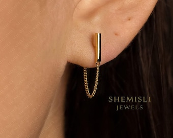 Może przedstawiać: Złoty kolczyk z pionowym pręcikiem i delikatnym łańcuszkiem. Kolczyk jest przymocowany do płatka ucha. Widoczny jest tekst "SHEMISLI JEWELS".