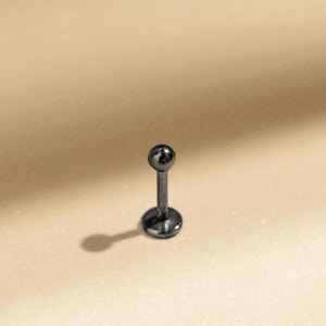 May include: A single, dark grey labret stud. The jewellery features a small, round ball at the top of a straight post, with a flat disc base. The stud is set against a neutral, light brown background.