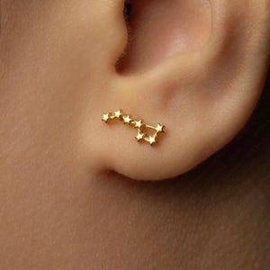 May include: A gold constellation ear climber earring. The earring is shaped like a series of connected stars, designed to be worn along the earlobe. The earring is shown on a person's ear.