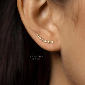May include: A gold ear climber earring featuring seven small, clear gemstones arranged in a curved line. The earring is worn on the ear, with the brand name "SHEMISLI JEWELS" visible below the ear.
