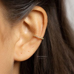 Peut inclure: Un ear cuff doré orné de petites pierres précieuses transparentes. Le bijou est porté sur le cartilage de l'oreille. L'image affiche également le nom de la marque "SHEMISLI JEWELS".