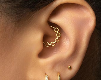May include: A gold daith hoop earring featuring a chain-link design inserted into the ear cartilage. Below, two additional gold hoop earrings are visible in the earlobe, one plain and one with small clear stones, plus a single gold stud earring.