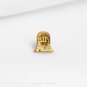 May include: A gold-coloured tombstone-shaped pin with the letters "RIP" engraved on the front. The pin has a textured surface and a rectangular base. The pin is a jewellery item.
