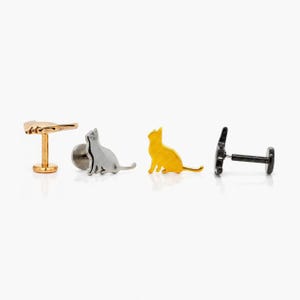 May include: Four pieces of jewellery are displayed against a white background. The pieces include a gold-coloured hand-shaped stud, a silver cat-shaped stud, a gold cat-shaped stud, and a black cat-shaped stud.