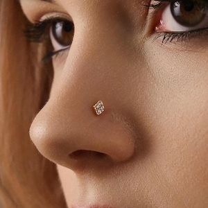 Peut inclure: Gros plan sur un nez orné d'un piercing de nez doré. Le bijou est de petite taille, en forme de losange, orné de plusieurs petites pierres transparentes. Le teint est clair, et l'accent est mis sur le nez et le bijou.