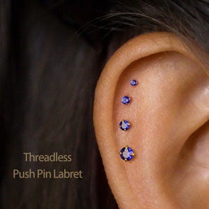 May include: An ear adorned with four threadless push pin labret earrings. Each earring has a bright blue gemstone set in gold-coloured metal. The text "Threadless Push Pin Labret" is displayed on the left side of the image.