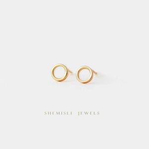 May include: Two small, gold-coloured circle stud earrings. The earrings have a simple design with a smooth, polished finish. They are displayed against a white background. The text "SHEMISLI JEWELS" is at the bottom of the image.