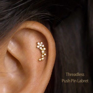 May include: A gold-coloured, curved labret stud featuring a cluster of clear, sparkling stones. The stud is crescent-shaped and set against the skin of an ear. Text on the image reads "Threadless Push Pin Labret."