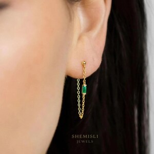 May include: A close-up of a gold chain earring featuring a small, rectangular emerald-green stone. The earring is attached to the ear with a small gold ball. The brand name "SHEMISLI JEWELS" is printed at the bottom.