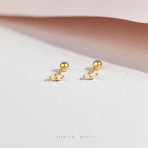 May include: A pair of gold-coloured stud earrings. Each earring has a small, round gold ball and a cluster of three small, white opal stones. The earrings are presented on a soft, light pink surface. The text "SHEMISLI JEWELS" is at the bottom.