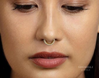 May include: A close-up of a face with a gold septum ring set with small, clear gemstones. The ring is horseshoe-shaped. The skin tone is light, and the lips are a deep red. The text "SHEMISLI JEWELS" is visible in the image.