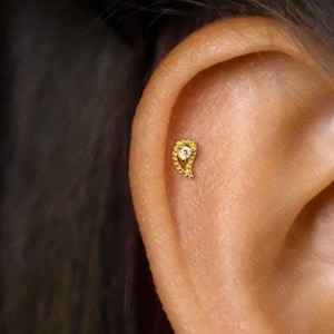 Peut inclure: Boucle d'oreille dorée en forme de paisley avec une petite pierre précieuse transparente au centre. La boucle d'oreille est placée contre la peau d'une oreille, mettant en valeur son design complexe et ses détails délicats.